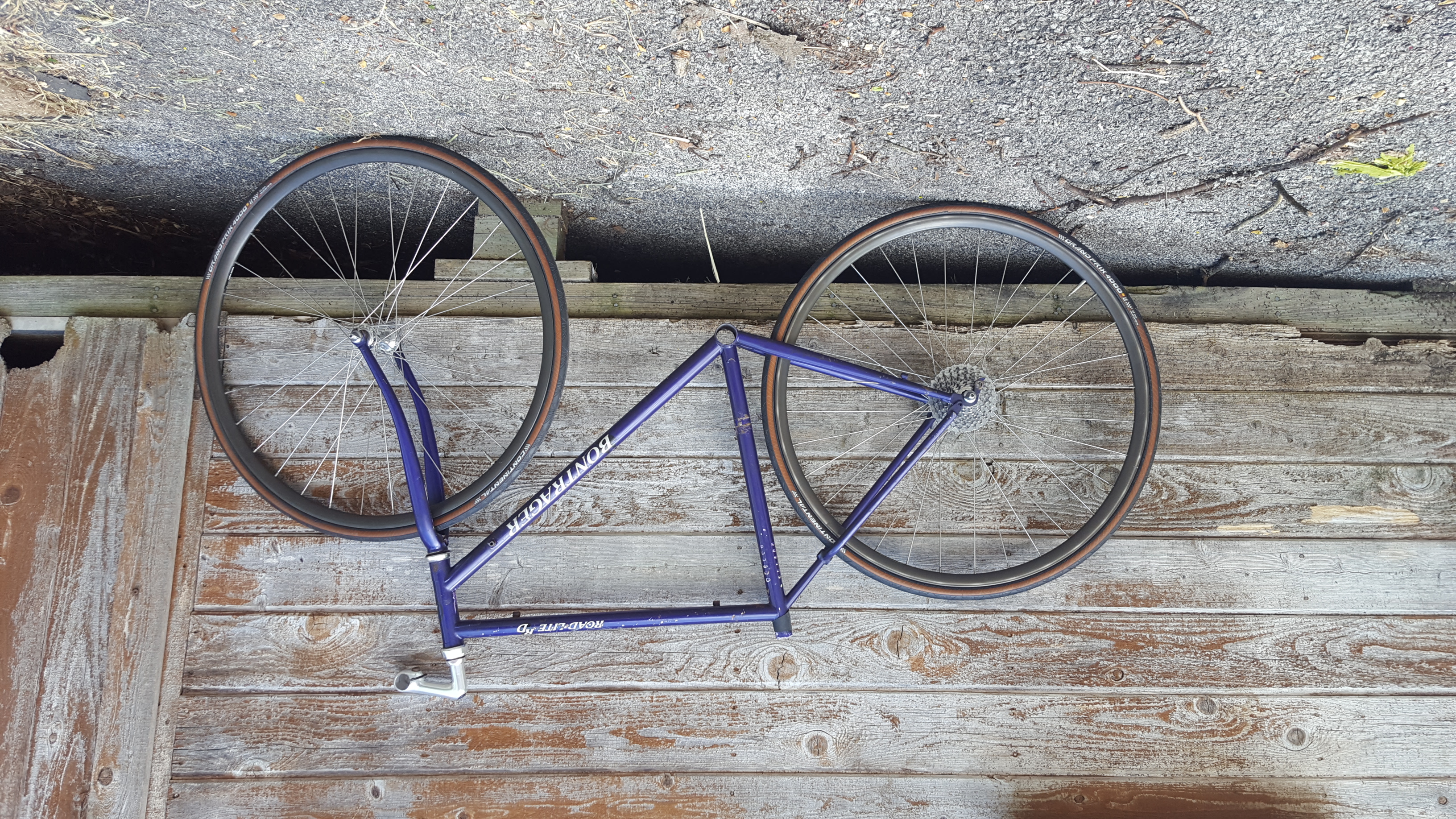 A scuffed and somewhat rusty deep-purple bicycle frame, with wheels and a stem, but otherwise no componentry posed in front of a shed.