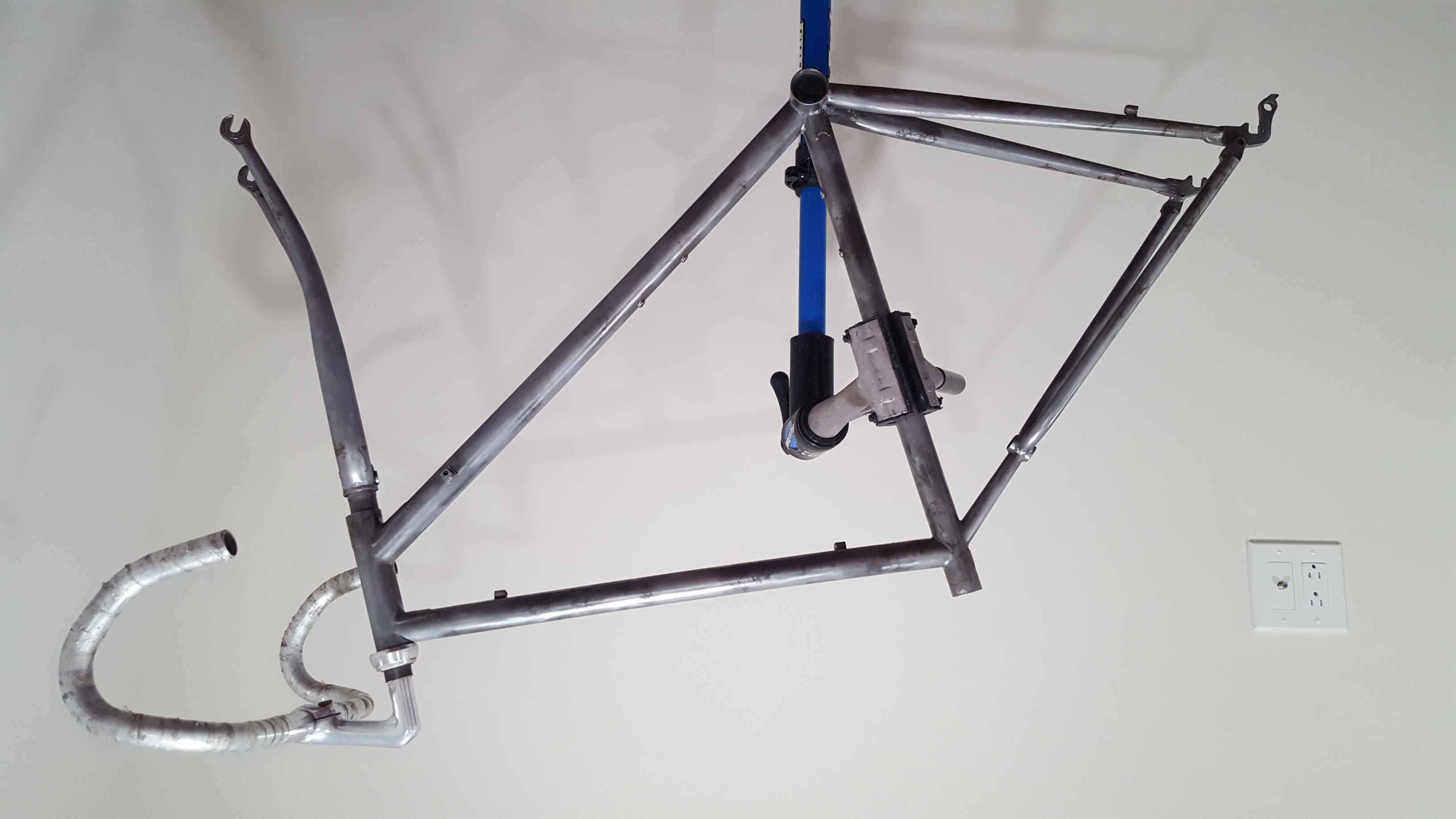 A bicycle frame, stripped of its paint, is in a blue bike stand, clamped by the seat-tube. The fork is inserted, with a stem and drop-handlebars. The background is a white wall.