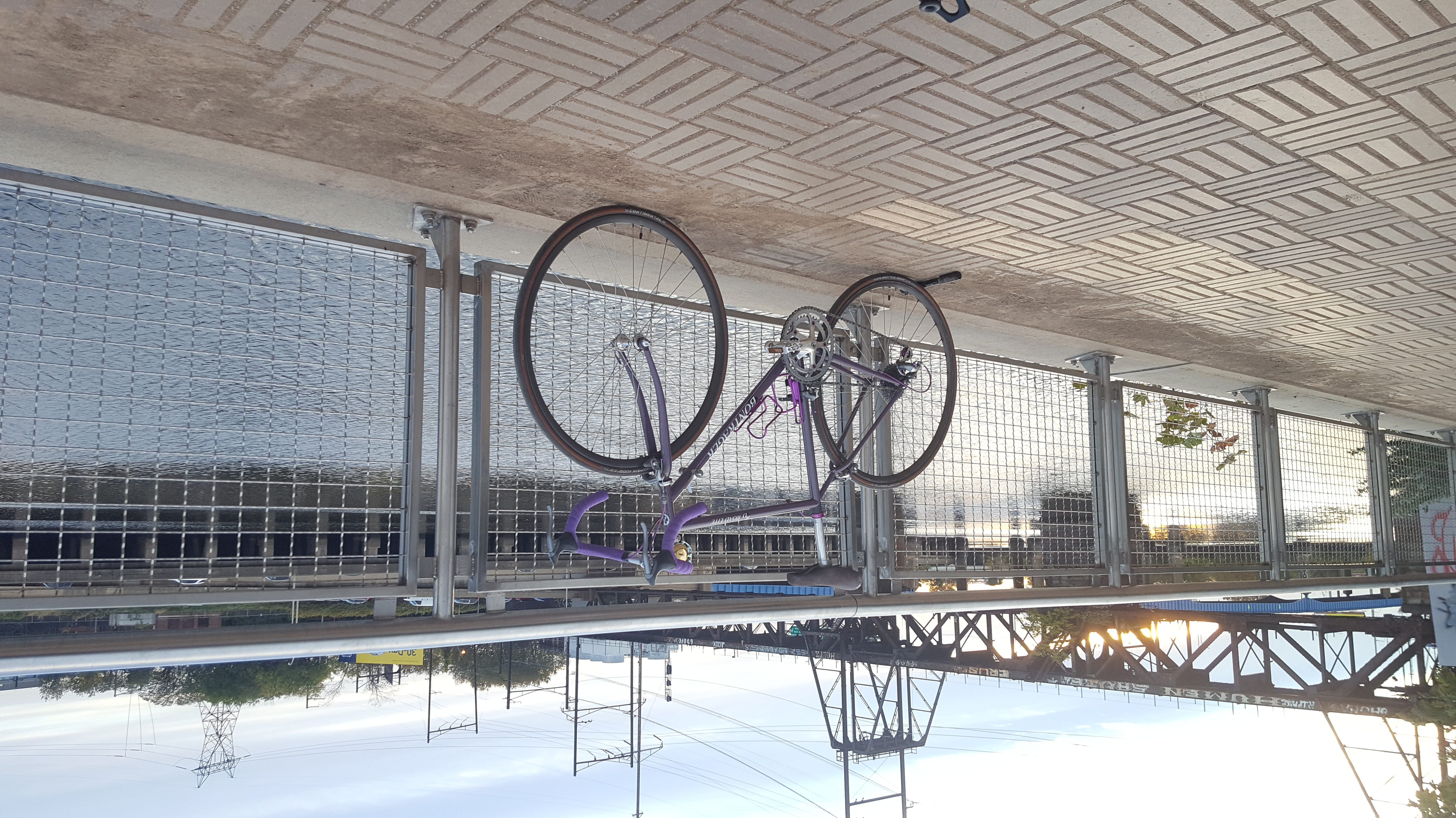 A purple bicycle posed on a fence in front of a river. A portable bike-pump can be seen under the rear tire, preventing it from rolling away. The camera has revolved around towards the front of the bike. In the background is a train bridge with the sun behind it.