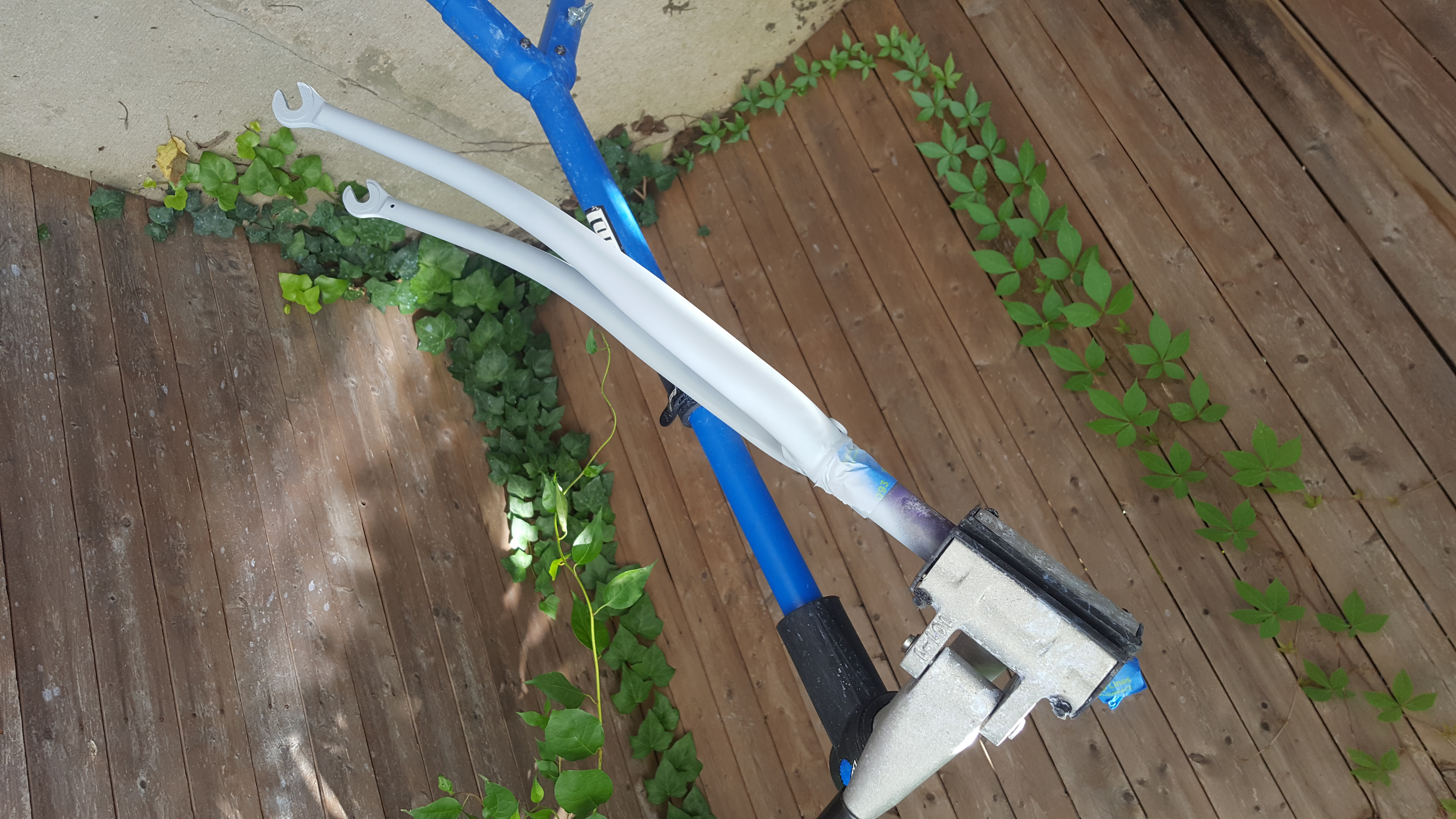 The bicycle fork is clamped by the steerer tube in a blue bike-stand. white-ish primer has recently been applied to the fork crown and stays. In the background is a fence with ivy on it.