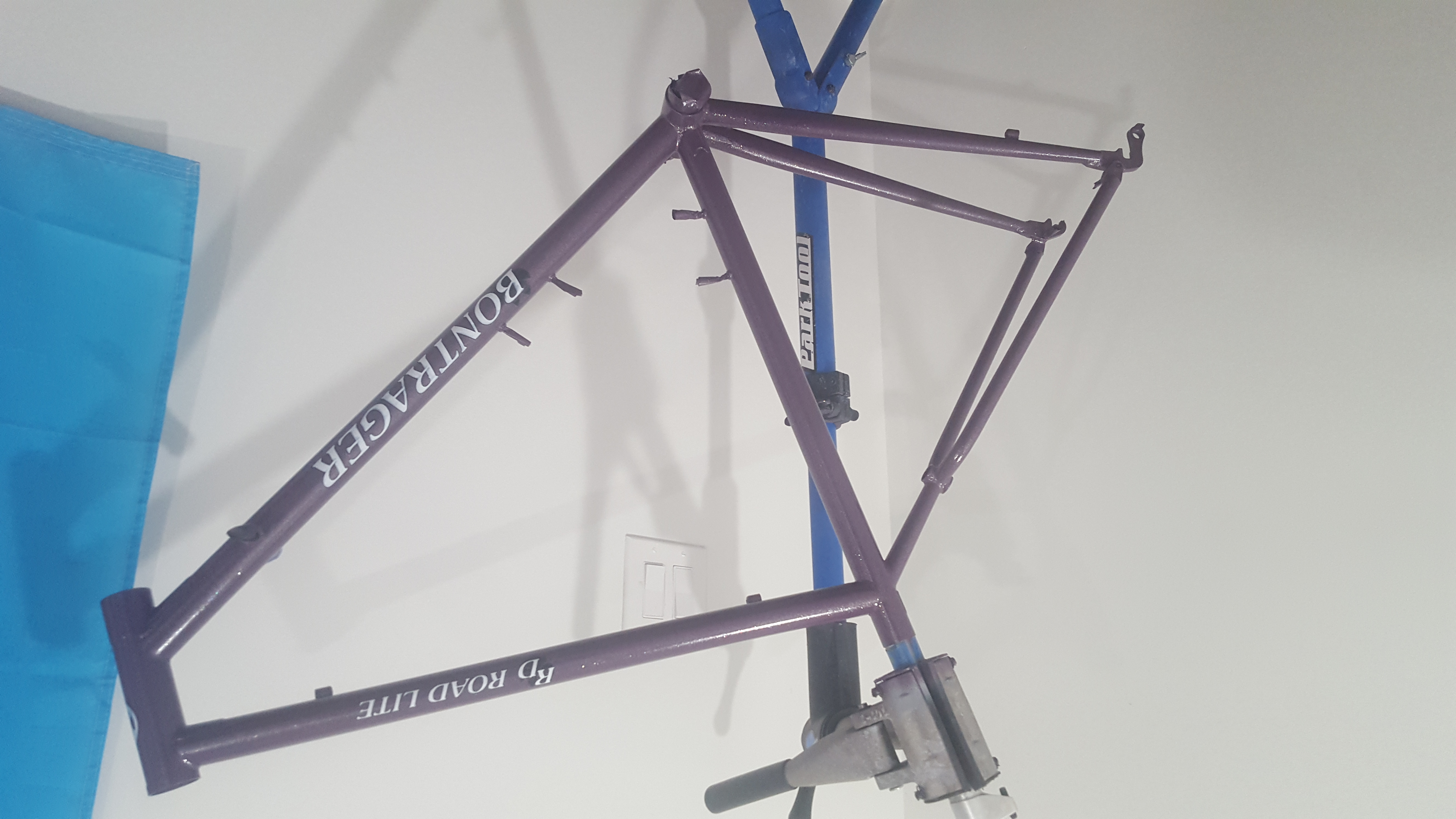 A recently painted purple bike frame is clamped by the seat-post in a blue bike-stand. Decals have recently been applied. 'Bontrager' appears on the down tube in all caps, and 'RoadLite' appears on the top-tube in small-caps. In the background is a white wall, with the blue-corner of a trans-pride flag appearing on the right side.