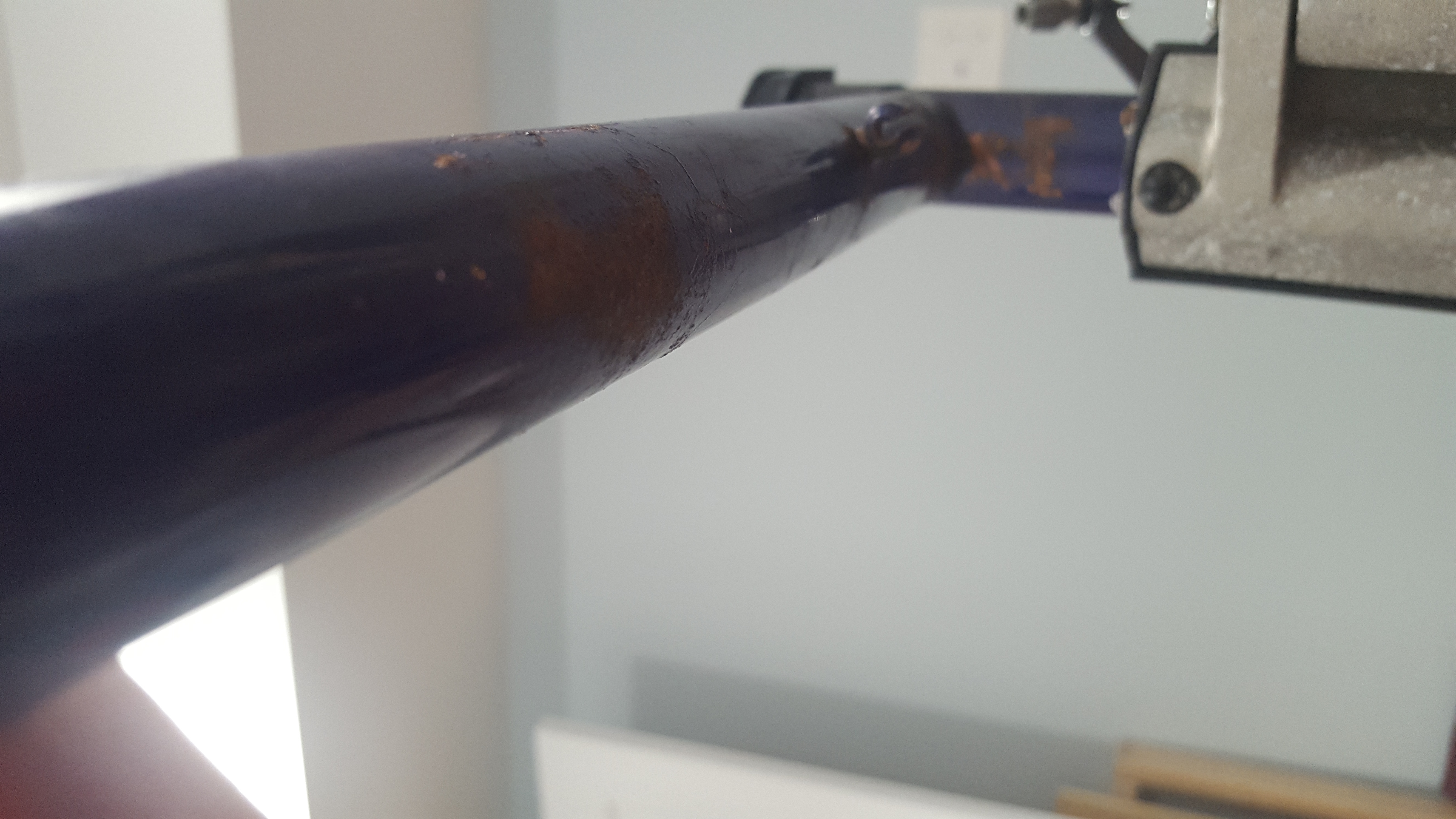A close up on the bicycle frame's top tube showing the joint with the seat tube. The seat tube is in the clamp of a bike stand. Rust is visible on the top tube and around the joint. In the background is a wall and ceiling.